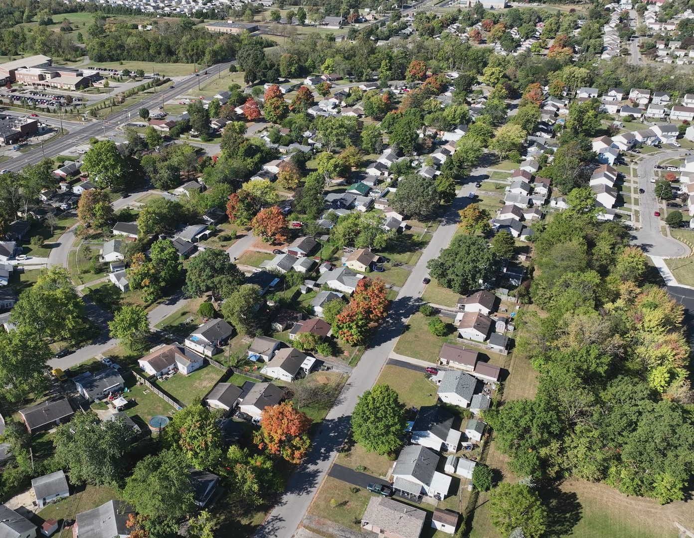 Aerial view of a suburban neighborhood with rows of houses, tree-lined streets, and patches of green lawns; businesses and roads are visible at the edge, with autumn foliage scattered throughout.