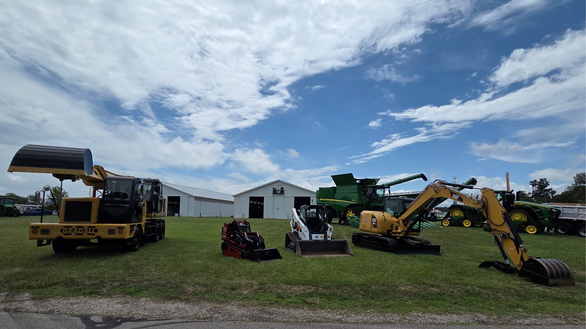 Several types of construction and farming equipment, including loaders, excavators, and combines, are lined up on grass in front of a white building under a partly cloudy sky.