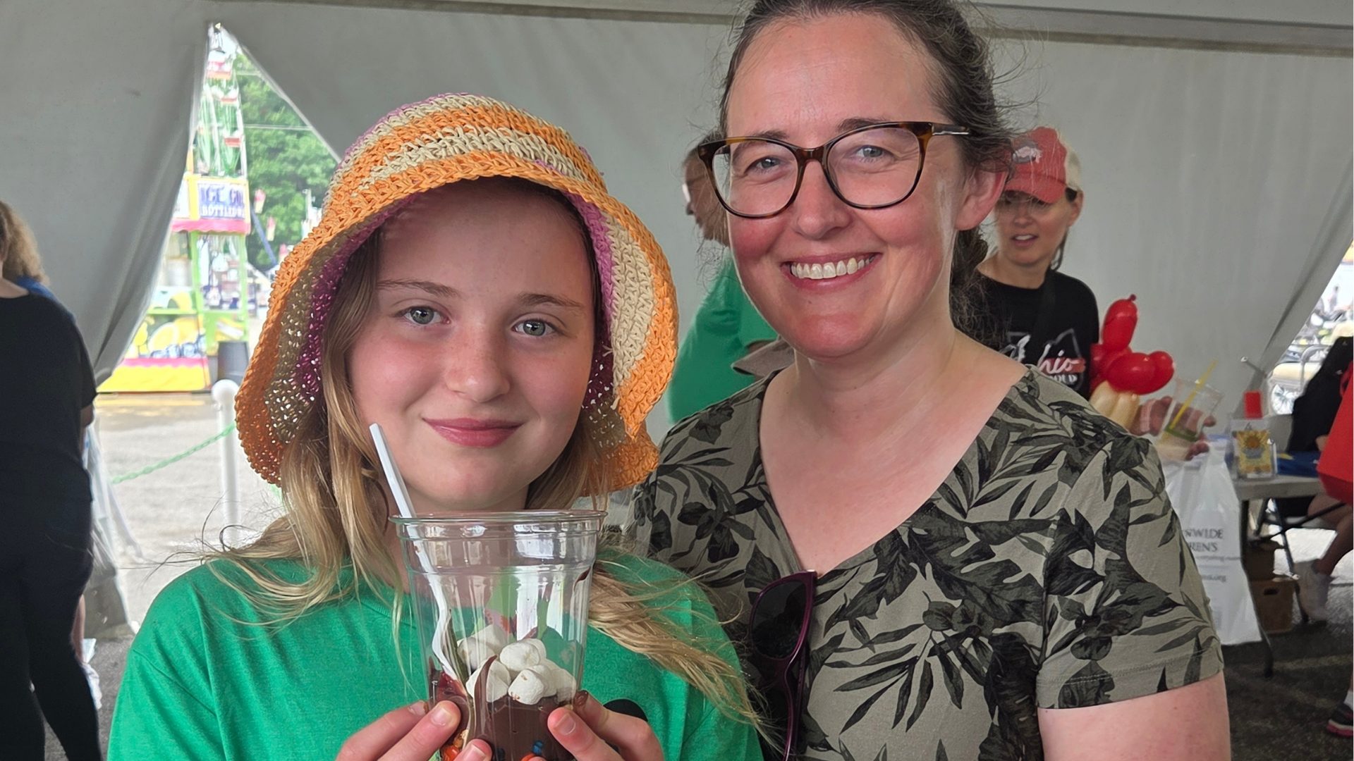 A smiling woman and a girl in a colorful hat hold a dessert in a clear cup under a tent at an outdoor event, with other people and festive decorations in the background.