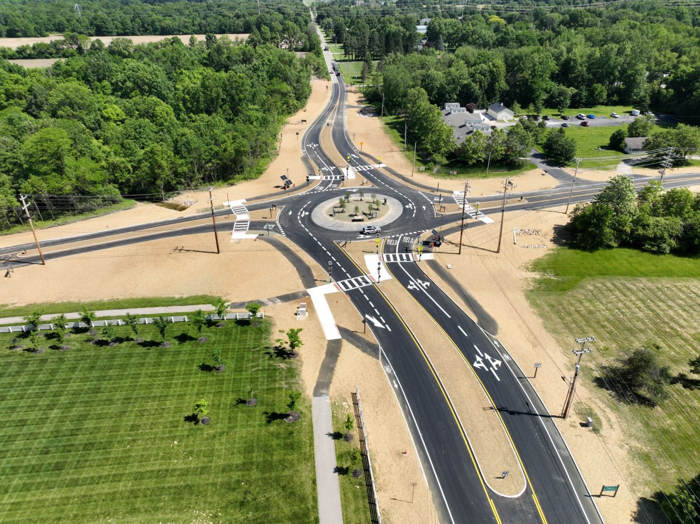 Aerial view of a newly constructed roundabout surrounded by green fields and trees, with multiple roads converging and clearly marked lanes, crosswalks, and traffic signs visible. Aerial view of a newly constructed roundabout surrounded by green fields and trees, with multiple roads converging and clearly marked lanes, crosswalks, and traffic signs visible.