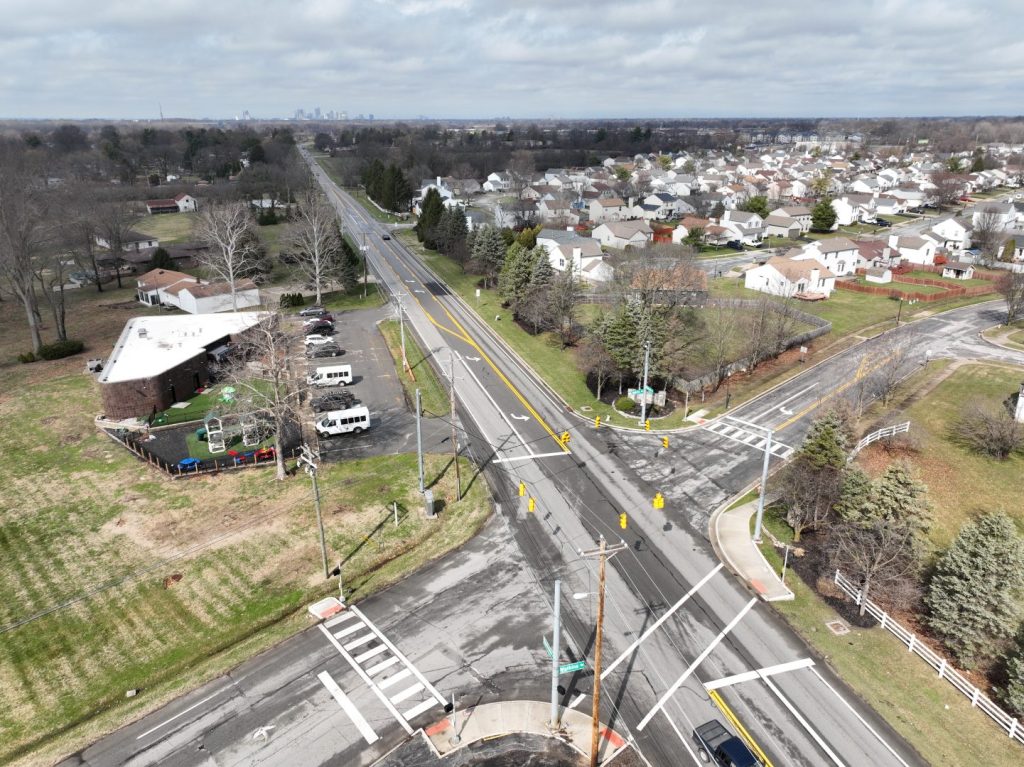 Aerial view of a suburban intersection with houses, a building with parked cars, grassy areas, and trees under a cloudy sky. Long roads extend toward the horizon where a distant city skyline is visible.