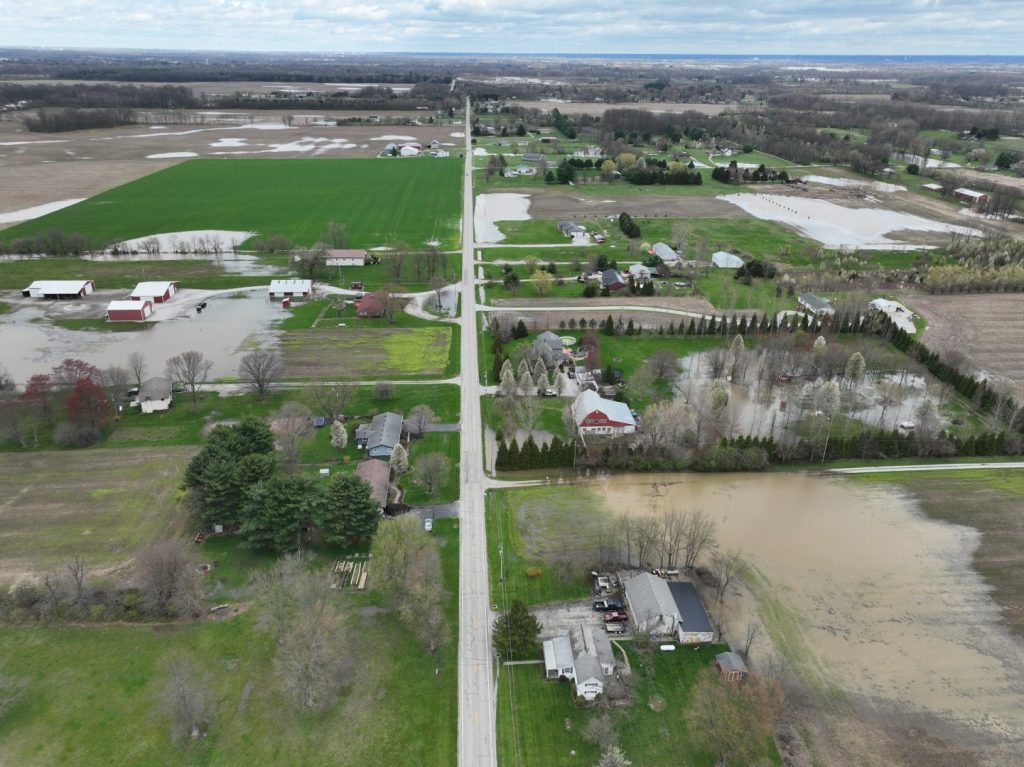 Aerial view of a rural area with flooded fields, roads, and homes. Some lawns and buildings are surrounded by water, while other areas remain dry. The sky is cloudy and trees are scattered throughout the landscape.