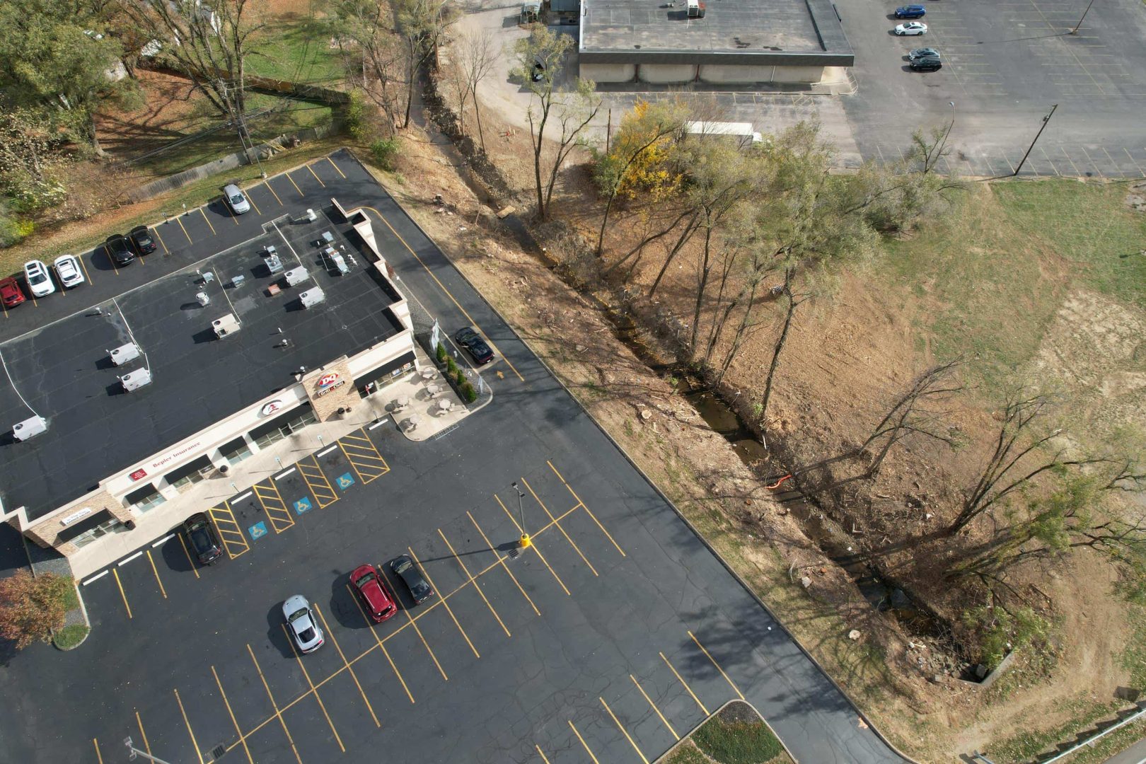 Aerial view of a small shopping plaza with mostly empty parking spaces, a few parked cars, and a tree-lined boundary next to a cleared grassy area. A larger building and more parking are in the background. Aerial view of a small shopping plaza with mostly empty parking spaces, a few parked cars, and a tree-lined boundary next to a cleared grassy area. A larger building and more parking are in the background.
