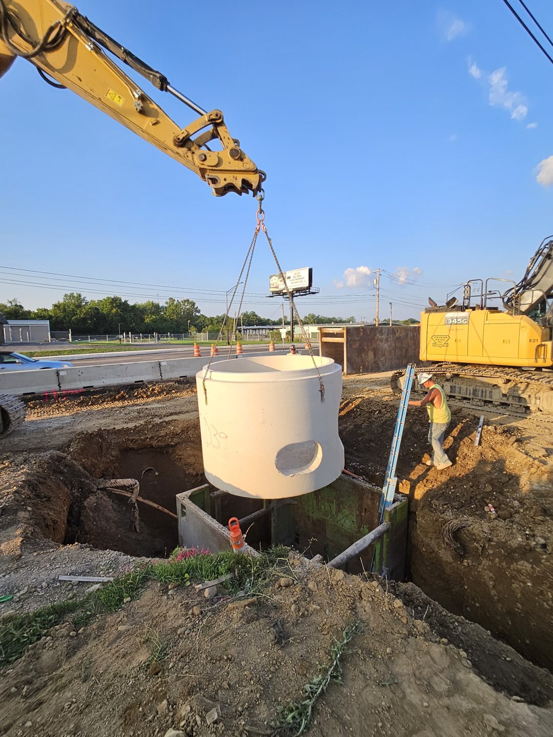 A construction site where a crane is lowering a large concrete cylinder into a pit, while a worker wearing a helmet stands nearby guiding the installation.