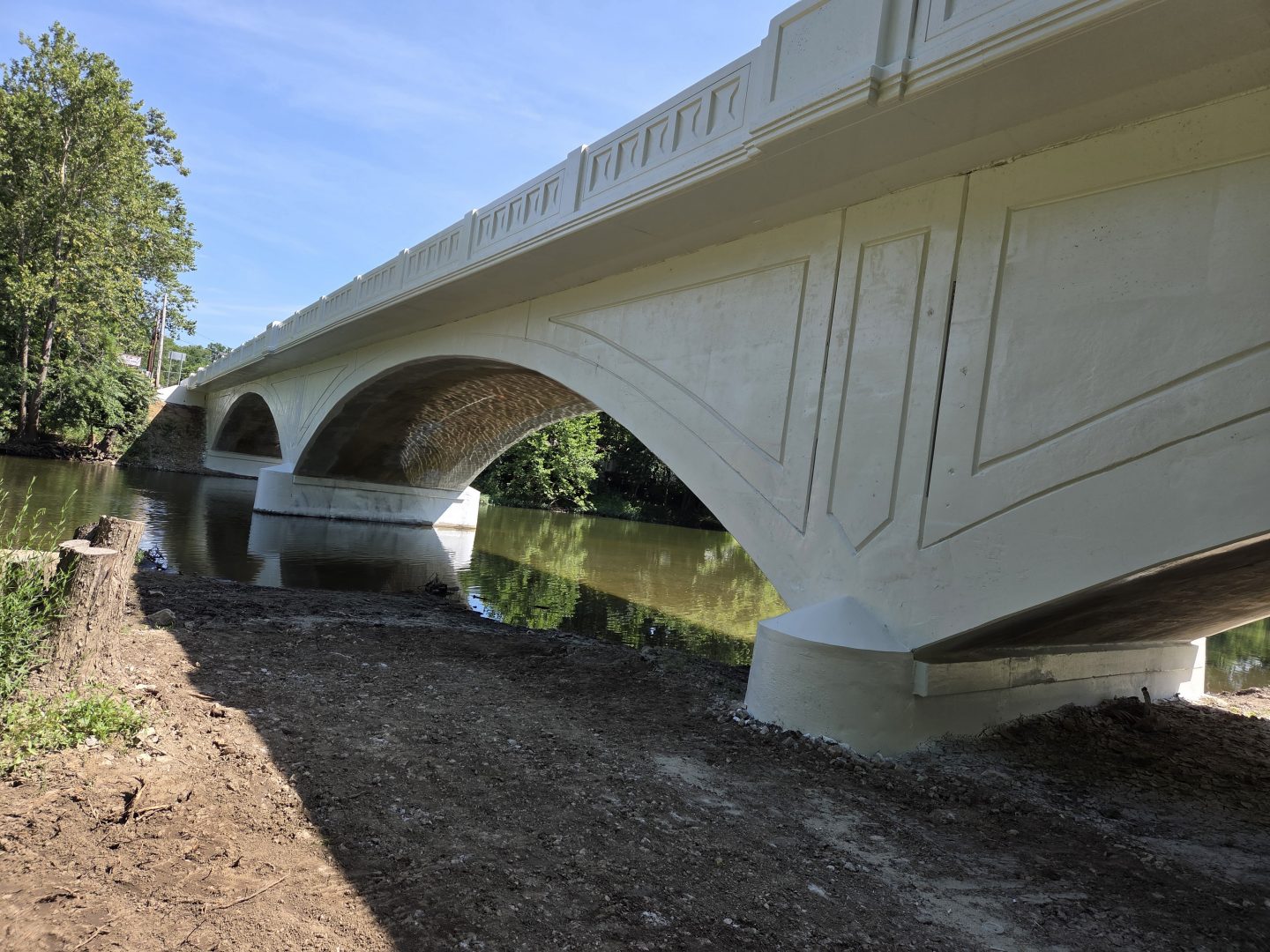 A white concrete bridge with arched supports spans over a calm river, surrounded by trees and blue sky, viewed from the shaded riverbank below.
