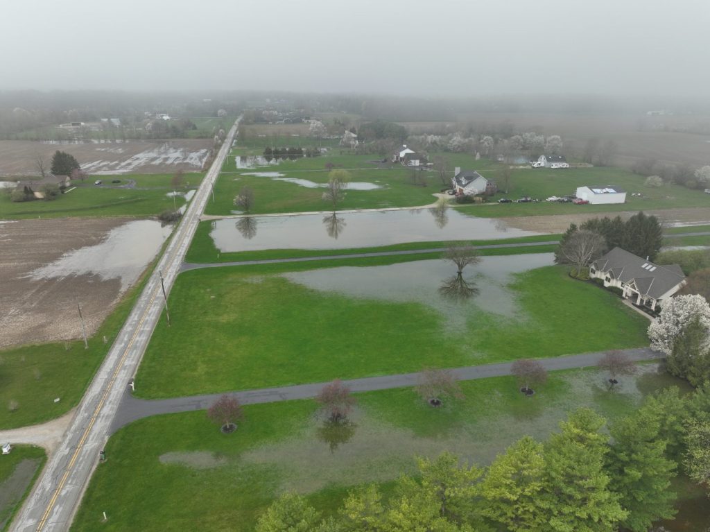 Aerial view of a rural neighborhood with flooded lawns and fields, cloudy sky, scattered houses, and water pooling along roads and grassy areas.