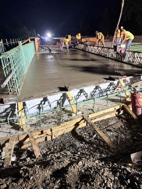 Construction workers at night pour and smooth fresh concrete on a bridge or roadway, with green rebar exposed along the edges and bright work lights illuminating the scene.