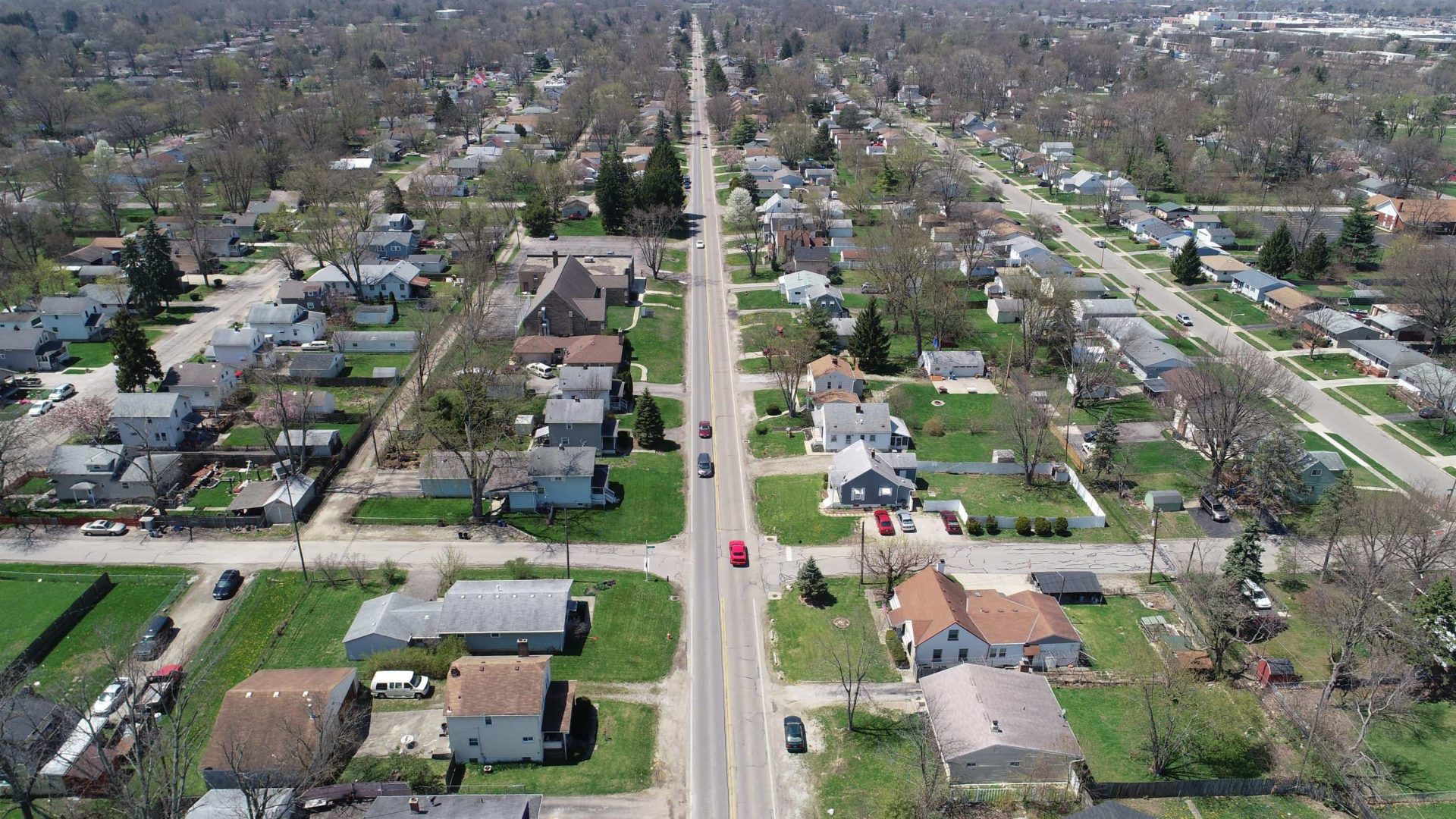 Aerial view of a suburban neighborhood with houses, lawns, and trees lining both sides of a straight road that runs through the center of the image. Several cars are parked along the street.
