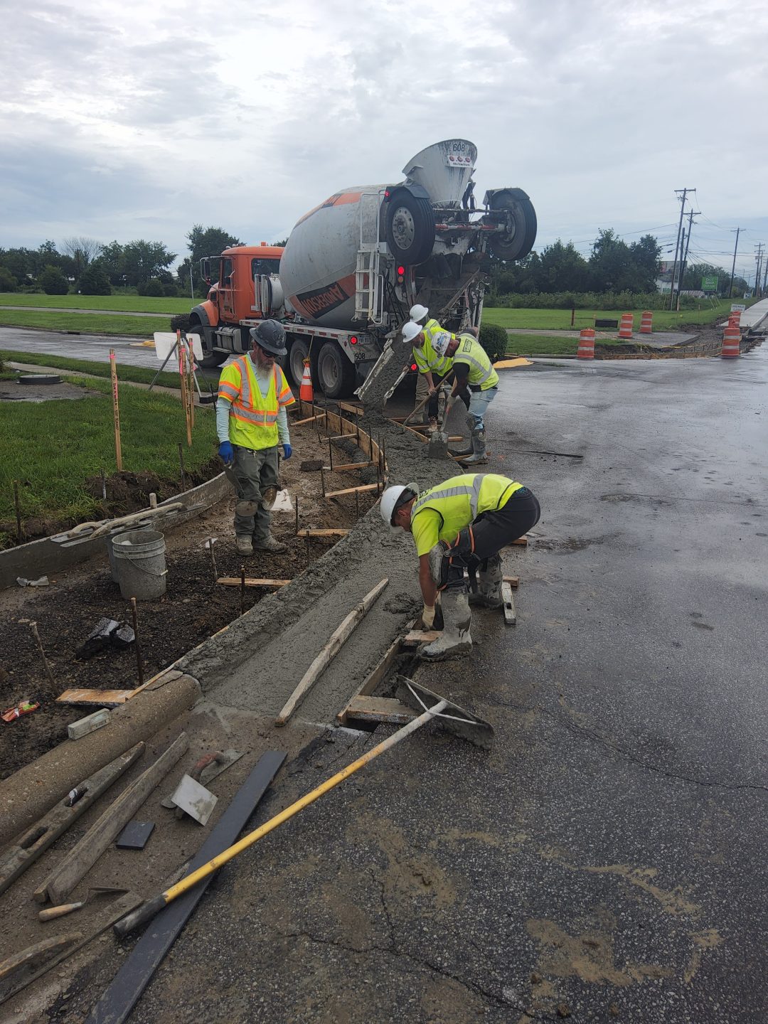 Three construction workers in high-visibility vests pour and smooth wet concrete for a new curb next to a cement mixer truck. Three construction workers in high-visibility vests pour and smooth wet concrete for a new curb next to a cement mixer truck.