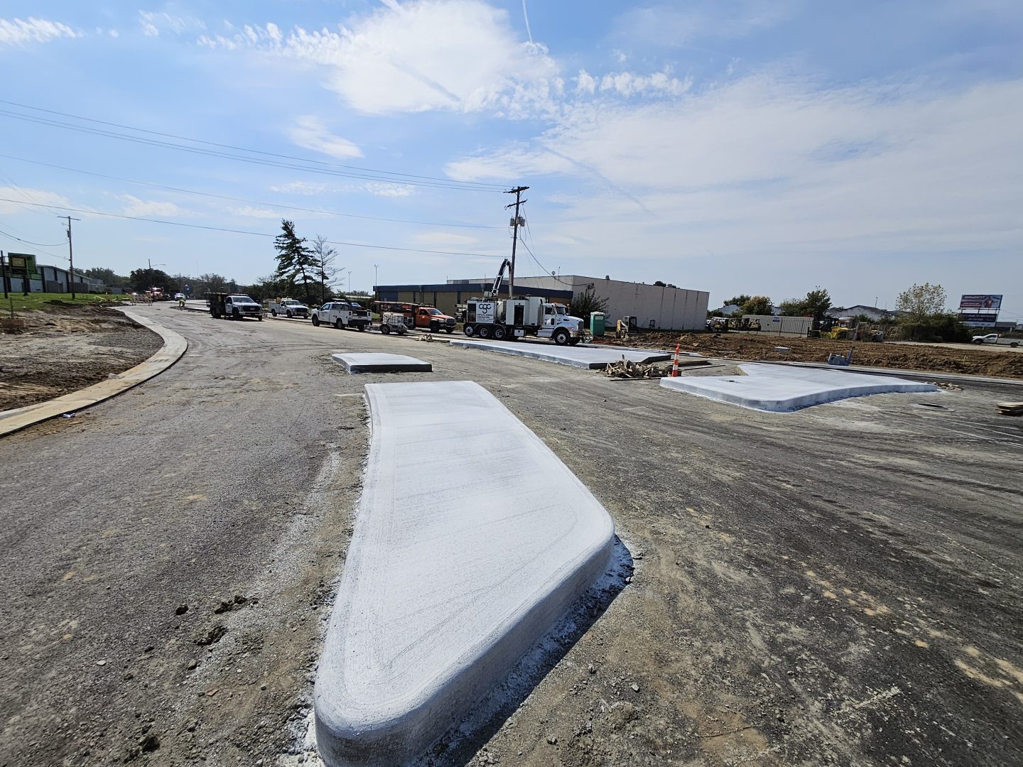 A construction site with newly poured concrete curbs on a gravel road, with work trucks and utility poles in the background. A construction site with newly poured concrete curbs on a gravel road, with work trucks and utility poles in the background.