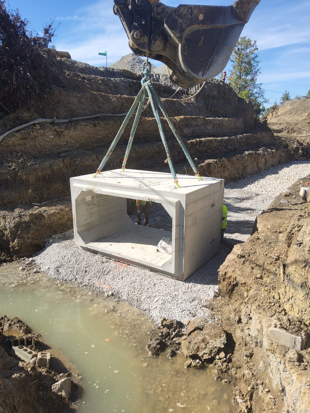 An excavator lowers a large, rectangular concrete culvert into a trench filled with gravel and water.