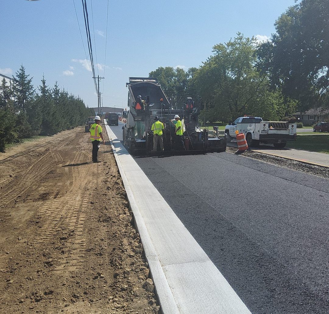 Construction workers use a paving machine to lay fresh asphalt on a road next to a newly installed concrete curb.